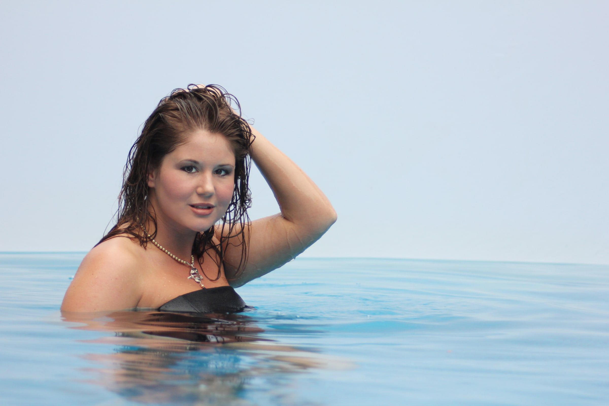 Model in the pool with one hand raised to her hair, necklace visible